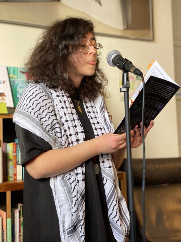 Summer wearing a keffiyeh, black dress, gold glasses, with her tongue sticking out slightly. She has brown hair and light skin. She stands in front of a microphone, holding a copy of I could die today and live again. 

